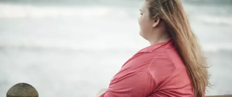 Photo of a young woman with her back turned. She's standing at a fence, gazing out at the ocean.